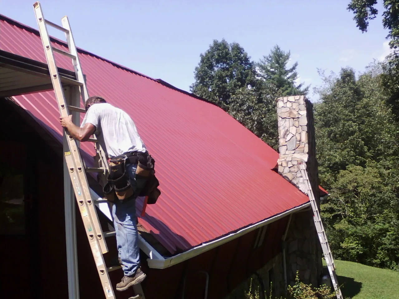 Roofer on a ladder beside a red metal roof and stone chimney — Soffit & Fascia Repair by Roofing Brothers in Van Wert